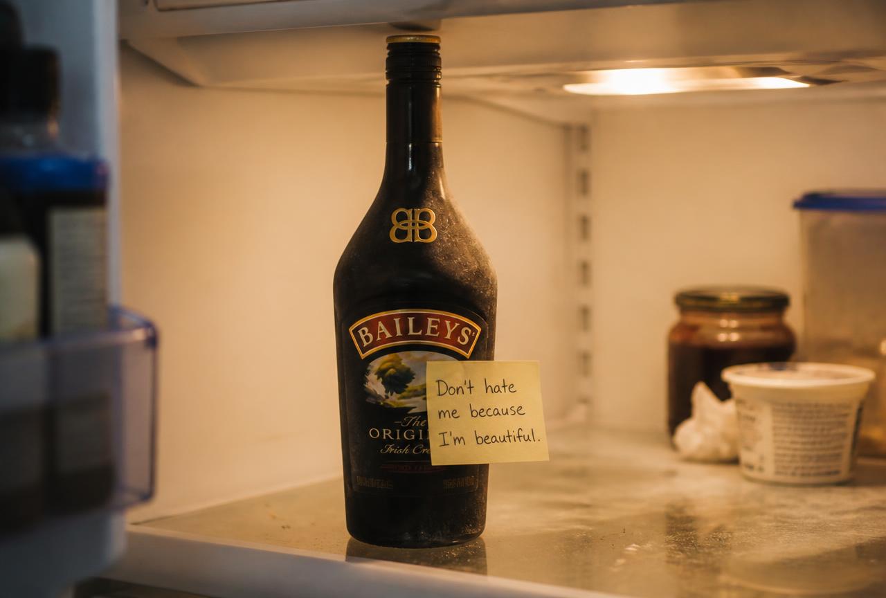 A dusty bottle of Baileys sitting alone on a fridge shelf with a handwritten sticky note on it